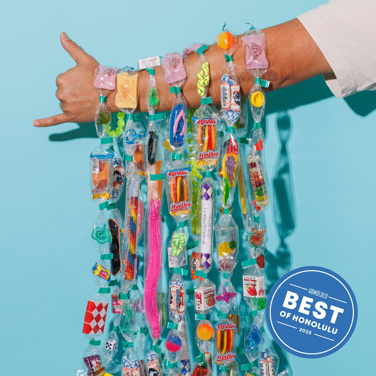 Person displaying colorful Trinket Lei on their arm with various small items on a blue background, with 'Best of Honolulu' badge.